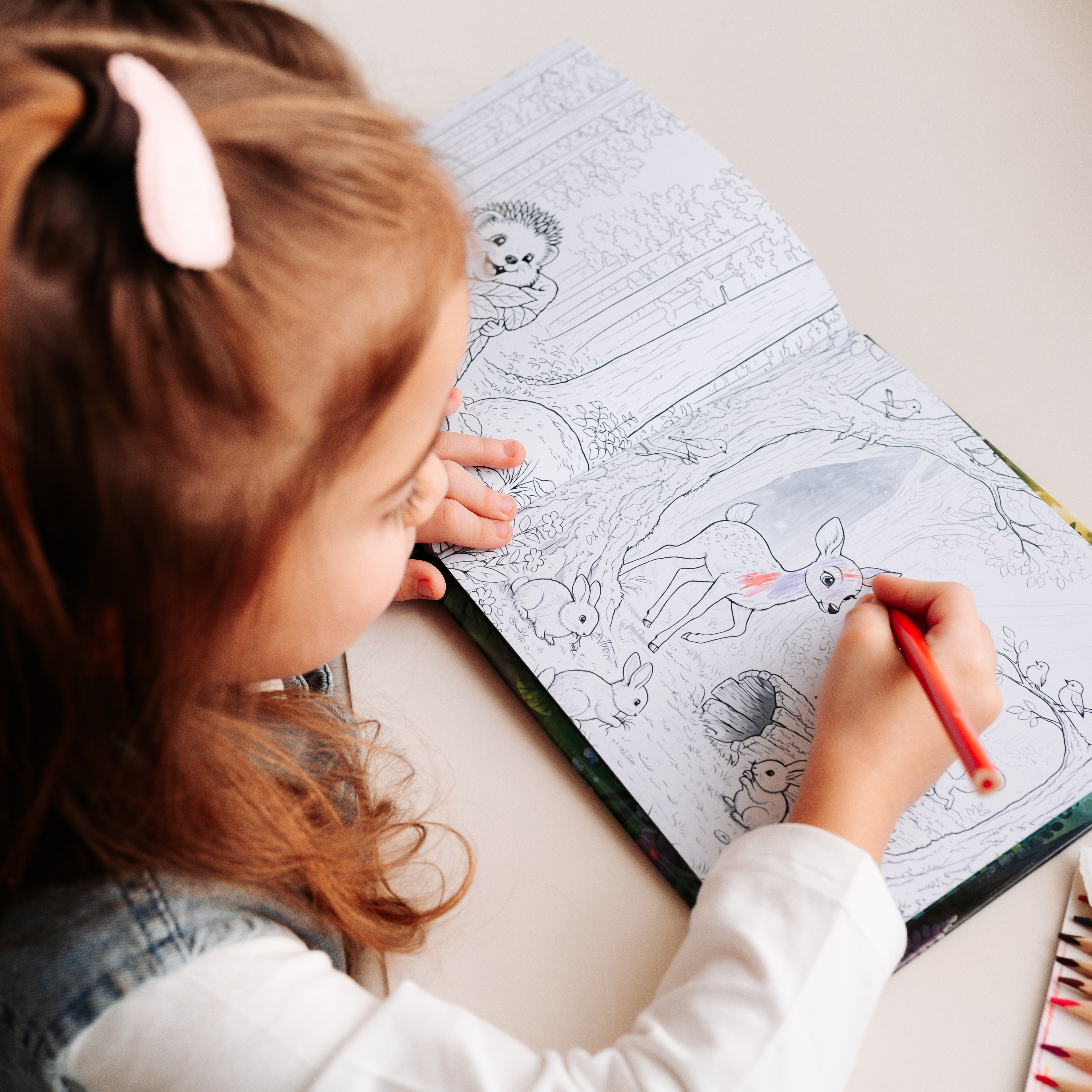 A young girl colors a black-and-white illustration in the "Child: Animal Helper" activity book, using a red pencil to fill in a detailed forest scene featuring a deer, rabbits, a hedgehog, and other woodland animals.