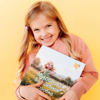 A smiling blonde girl in a pink sweatshirt proudly holds her personalized children's book titled "Milana and the Magic Box" featuring a cartoon girl running through a sunny meadow on the cover.