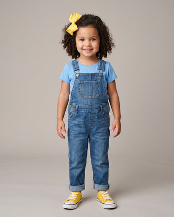 A young child with curly dark hair wearing a yellow bow, a light blue t-shirt, blue denim overalls, and yellow sneakers, standing against a neutral gray studio background.