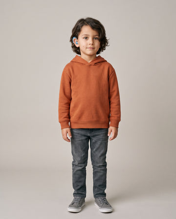 A young boy with dark curly hair stands facing forward in a studio, wearing a rust-orange pullover hoodie, dark gray jeans, and gray sneakers against a light gray background.