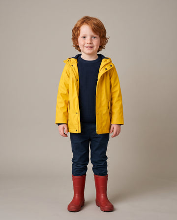 A young child with red hair stands against a plain beige studio background wearing a bright yellow hooded rain jacket over a dark navy ribbed sweater, navy pants, and red rain boots.