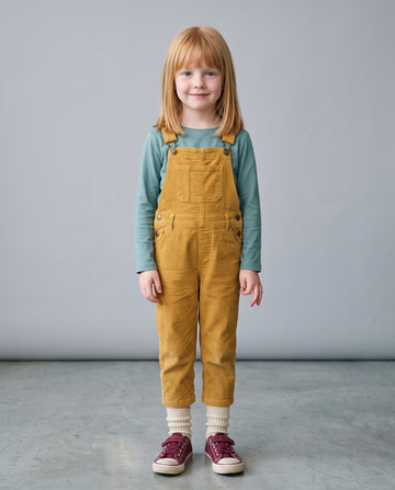 A young girl with strawberry-blonde hair stands wearing mustard yellow corduroy bib overalls over a sage green long-sleeve shirt, cream knit socks, and burgundy sneakers, against a neutral gray studio background.