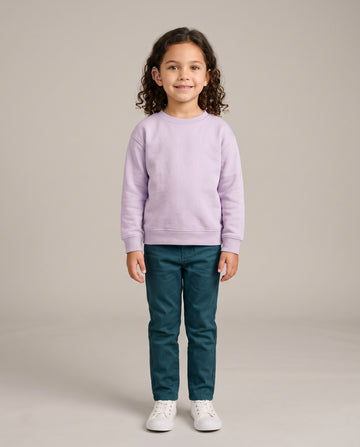 A young girl with curly brown hair stands against a neutral beige studio background, wearing a light lavender crewneck sweatshirt, dark teal pants, and white sneakers.