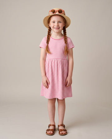 A young girl with braided hair stands against a plain beige studio background wearing a pink and white striped short-sleeve dress, a straw sun hat with sunglasses, and brown sandals.