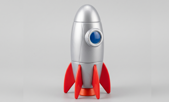 A retro-style toy rocket with a metallic silver body, red fins, and a blue circular window, standing upright on a light gray background.